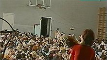 Hundreds of hostages packed into the school gym with wired explosives attached to the basketball hoop (a frame from the Aushev tape) Beslan School Terror - terrorist 4 - Sept 1, 2004.jpg