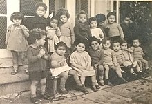 8 17 mixed race children sitting and standing outside the Holnicote House building in England in the 1940s. The children are wearing dresses or shorts.