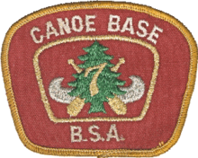 Patch with a red background and a gold border; the text Canoe Base BSA in the border; in the center a silver canoe superimposed with crossed paddles, a green pine tree and the number 7 in gold