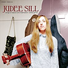 A photo of Sill holding a guitar, standing in front of a coat rack, smiling