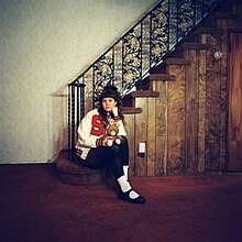 Pouty seated at the bottom of a staircase, holding a teddy bear and wearing a letterman's jacket