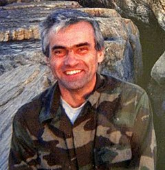 A smiling man with salt-and-pepper hair, slightly disheveled, wearing a coat in a camouflage pattern with rocks behind him.