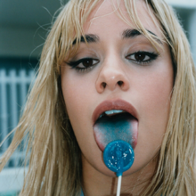 A young woman with long bleached hair and make-up looks at the camera with her mouth open and tongue blue from licking a lollipop.