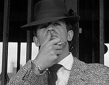 Black-and-white image of a man seen from mid-chest up, wearing a fedora and a jacket with a houndstooth-like pattern. He holds a cigarette between the middle and index fingers of his left hand and strokes his upper lip with his thumb. He stands in front of what appears to be a mirrored doorway.
