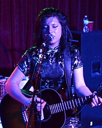 Chloe Chaidez se apresentando no palco;  cantando em um microfone e tocando uma guitarra