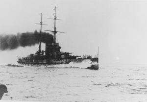 Een groot slagschip stoomt door het water.  Water breekt tegen de boeg terwijl zware donkere rook uit de twee trechters van het schip komt.