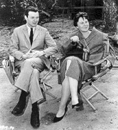 Une photographie en noir et blanc d'Alan J. Pakula assis à côté de Harper Lee dans les fauteuils du réalisateur en train de regarder le tournage de To Kill a Mockingbird