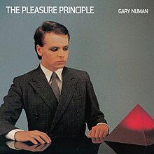 Gary Numan (seen wearing a business suit) looks at a glowing purple Perspex pyramid that is on a black reflective table.