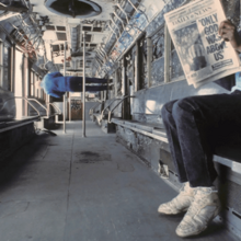 The inside of an abandoned subway car A person is seen sitting and holding a newspaper with the headline ONLY GOD WAS ABOVE US Farther down an out-of-focus person is seen standing on the side of the car with their back towards the camera