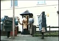 Three people dressed in cardboard costumes as a camcorder, microphone and clapperboard respectively, meet a fourth person with a large cardboard head and arms (seen here in the centre exiting the house) in the Fukme 99 video. Juxtapozed-With-U-Fukme99-SFA-Screenshot.jpg