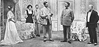 scene from a stage play with six characters - two women and three men - in Edwardian dress grouped in a luxurious drawing room