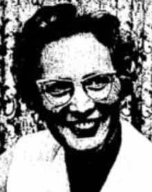 A black-and-white photograph of a smiling white woman with coiffed curly short hair and glasses