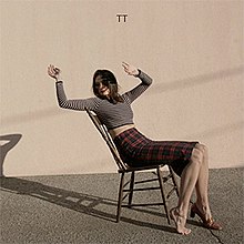 An image of a woman leaning backwards on a wooden chair in front of a beige-colored wall. Her arms are suspended; her right foot is missing a shoe. Uppercase text in a black serif font above her reads "TT".