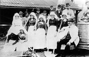 Grande grupo de homens com chapéus-coco e mulheres com gorros grandes, posando com ferramentas