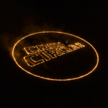 The title "Crop Circle 2" inside of a circle against a black background, with the text outlined in orange as if on fire in a field, with some smoke visible
