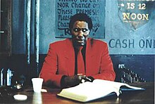 A middle-aged black man in a bright red suit sits at a dark brown desk against a backdrop of a wall painted in various dull shades of blue. His bejeweled hands are folded, and he is frowning with eyes focused off-camera to his left.