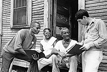 Black and white photograph of a black woman and a black and a white male poll worker explaining voting registration to a seated black man.