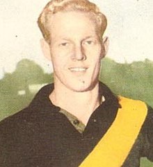 Egy szőke férfi elmosolyodik, miközben fekete, sárga szárnyú futballugrót visel