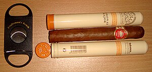 A double guillotine-style cutter, used for cutting the tip off a cigar, next to two hand-rolled H. Upmann Coronas Major cigars, one inside its storage tube and one outside. H.Upmann Coronas Major.jpg