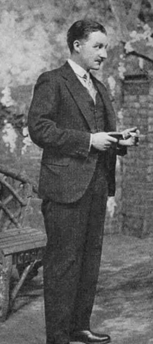 Tallish white man in lounge suit; he has a full head of neat, dark hair