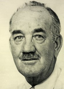 Black and white photo of a man with balding black and grey hair and a small moustache, wearing a white dress shirt and a necktie