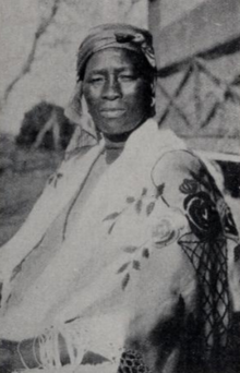 A photograph of a seated African woman wearing a turban and traditional dress in front of a building out of doors
