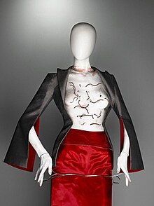 Look 64, corset made from translucent plastic with real worms embedded within, worn with a red-lined grey jacket and red silk skirt. At the hips, an item of unconventional silver jewellery which creator Shaun Leane calls a "stag piece". The worm corset.jpg