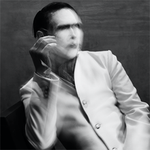 Image of a man wearing heavy make-up and a white jacket, who is sitting on a black chair and a grey-painted backdrop is visible behind him. The man's face is partially obscured by blurring, as is his right hand, which is being held upright.