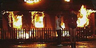 Rihanna and Eminem are standing in front of a burning house in a dark setting.