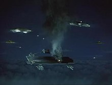 An airborne aircraft carrier, hovering at high altitude, is under missile attack from a group of flat, circular alien spacecraft and emitting fire and smoke. The setting is night.