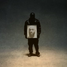 Ty Dolla $ign (a co-author of the album) is wearing a leather jacket and a mask. He is standing in the center of a brown background while holding a picture of his brother "Big TC".