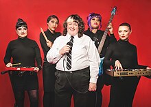 Fatty Cakes and the Puff Pastries standing in front of a red back drop. Crow, Audrey, Vishinna, and Stacey are wearing all Black with silver accents with a playful but serious look. Amber is standing in the center with a white dress shirt and black pants and a black tie, holding a microphone and smiling.