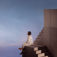 Capaldi sitting on a landing between flights of stairs, facing away from the camera towards a blue sky background