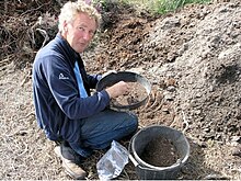 Tilley, an older white man with grey hair, kneeling while sieving a soil sample.