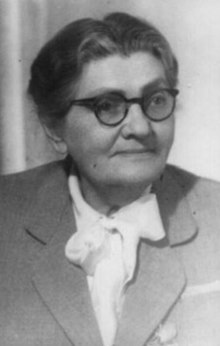 Black and white photograph of the bust of a woman wearing glasses, a white blouse with a large bow at the neck, and a double-breasted suit jacket.