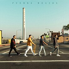 Four men walking on a sidewalk, with the Santa Fe Smokestack and a hotel seen in the background.