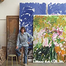 A photograph of Joan Mitchell in her painting studio in Vétheuil, France, with large abstract paintings behind her