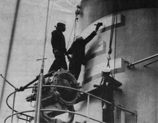 Crewmembers paint a gold "E" on the stack of the USS Rankin