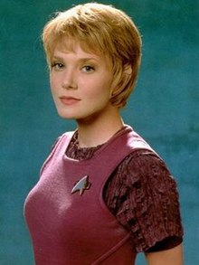 Uma fotografia de uma jovem com cabelo loiro curto, vestindo uma roupa marrom, com um broche de Star Trek anexado.