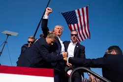 Trump raising his fist and shouting, with blood on his ear, while being carried by Secret Service agents. There is an American flag nearby in the background.