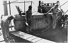 La tourelle d'un sous-marin à quai.  Quatre hommes se tiennent autour de la tourelle et une planche de passerelle a été posée à côté.  On peut également voir l'écoutille ouverte du sous-marin et diverses cordes amarrant le bateau.