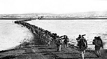 A long orderly line of heavily ladened soldiers marching in pairs away from the camera down a road across an open expanse.