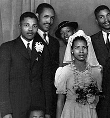 Una fotografía en blanco y negro de un hombre negro y una mujer negra, uno al lado del otro.  Él lleva un traje, ella lleva un vestido blanco.