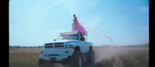 Charli XCX is seen in a pink fluffy ballgown while standing on top on a white monster truck. Charli XCX, White Mercedes.png