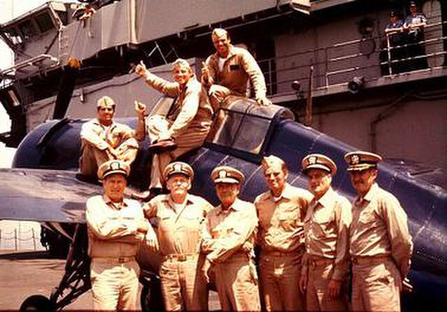 Nine members of the cast pose with a Grumman F4F Wildcat fighter on the flight deck of USS Lexington