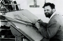 Een bebaarde man van midden dertig, gekleed in een hemd en een wollen trui, zit aan een tekentafel.  Zijn potlood ligt in zijn rechterhand en hij lacht links van de camera.