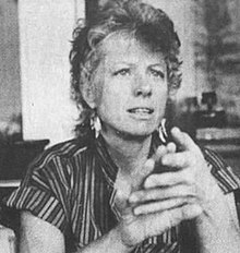 Black and white photograph of the head and shoulders of a woman in a striped blouse with short hair and hoop earrings seated with her elbows on an unseen surface and her hands raised and crossed at her neck-level