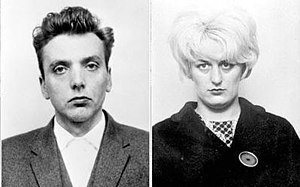 An unsmiling blonde woman with a short bouffant hairstyle, wearing a dark coat. To her left is a scowling man with dark, combed-back hair. Both are staring directly at the camera.