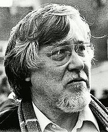 Three-quarter profile, outdoor shot of a bearded and bespectacled man, with strong features and almost-shoulder-length greying hair, in casual dress