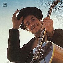 Дилан смотрит в камеру, держа гитару, улыбаясь и снимая кепку.